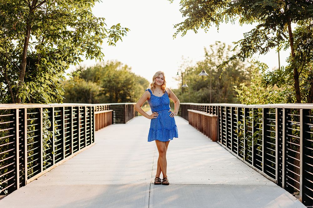 A person in a blue dress poses on a modern pedestrian bridge surrounded by green trees in sunlight.