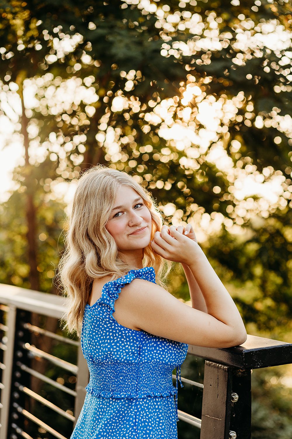 A person in a blue polka dot dress poses outdoors at sunset with golden bokeh light filtering through trees.