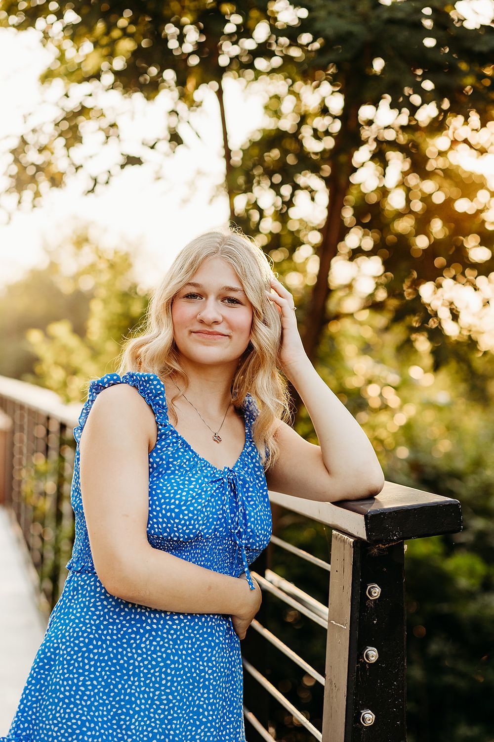 A person in a blue polka dot dress poses outdoors at sunset, leaning against a wooden railing with golden bokeh lighting.