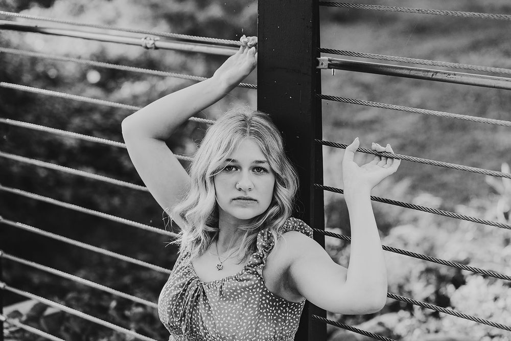 Black and white portrait of person in polka dot dress posing against metal railings in outdoor setting.