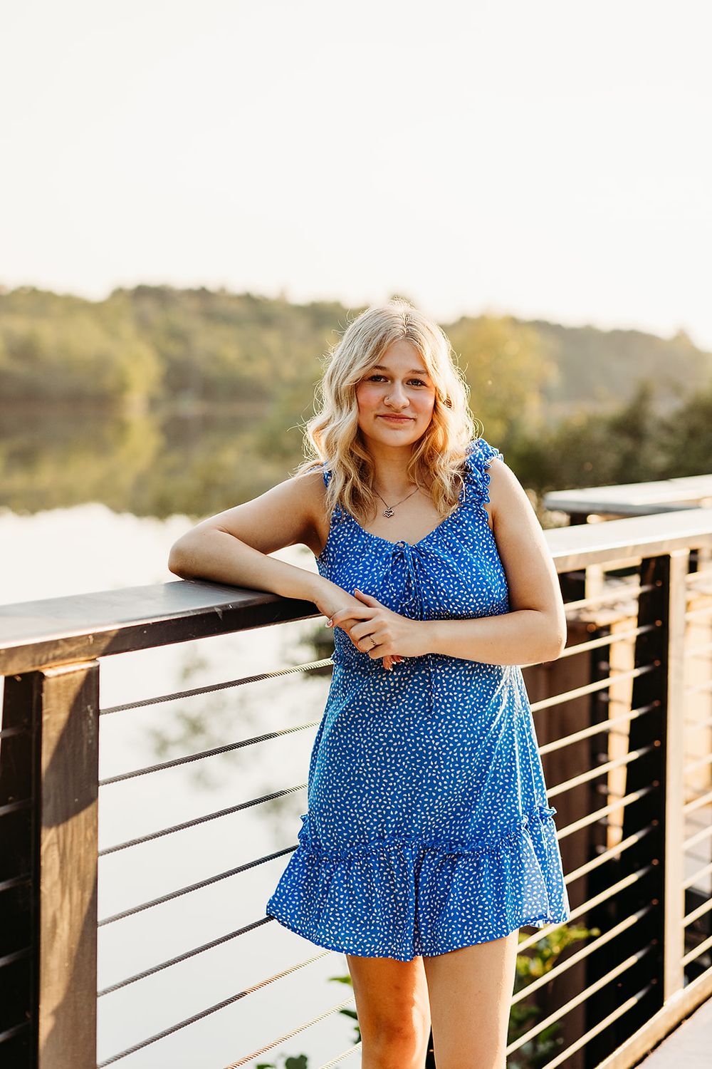Person in blue polka dot dress smiles while standing on wooden deck overlooking scenic landscape at sunset.