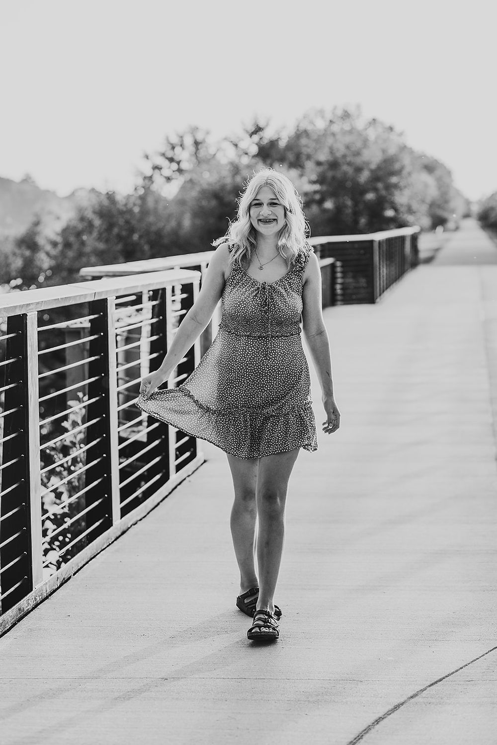 Black and white portrait of someone walking on a bridge wearing a flowy polka dot sundress on a sunny day.