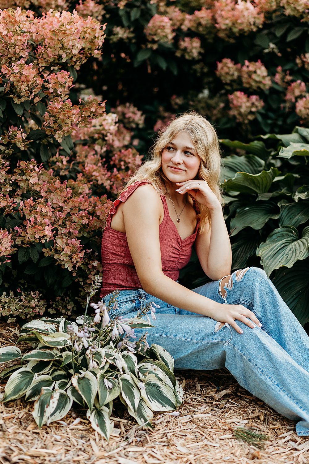 Outdoor portrait featuring casual red tank top and jeans against flowering pink blooms and greenery.