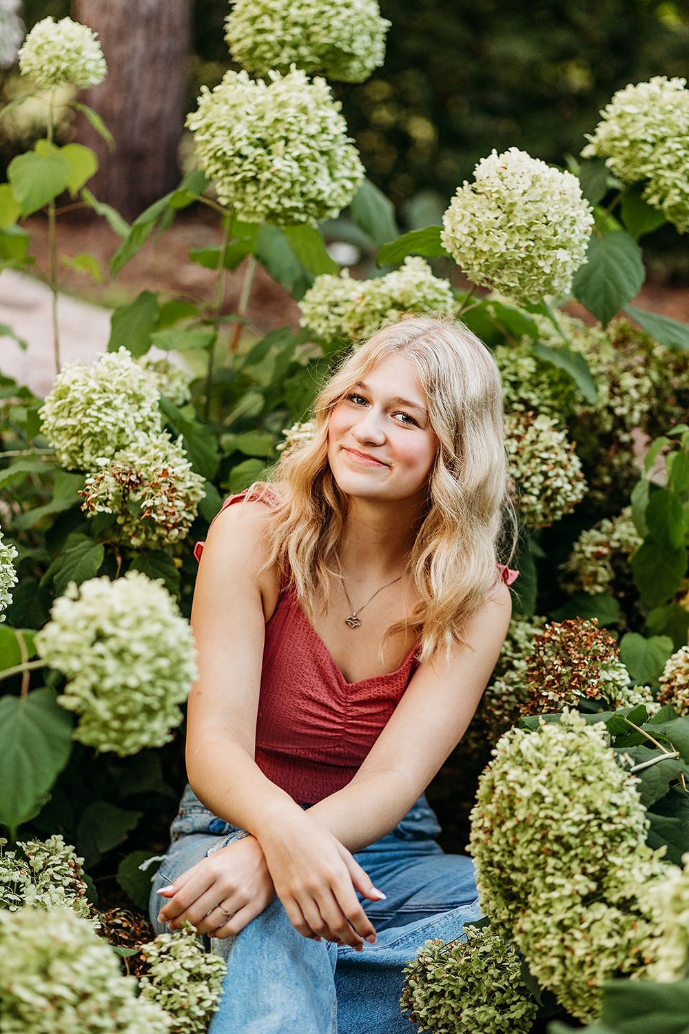A candid portrait in a garden with lush green hydrangea blooms, featuring someone in a burgundy top and jeans.