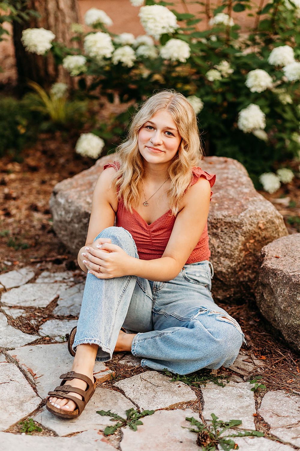 A person in a red tank top and jeans sits casually on stone pavers with white hydrangea blooms in the background.