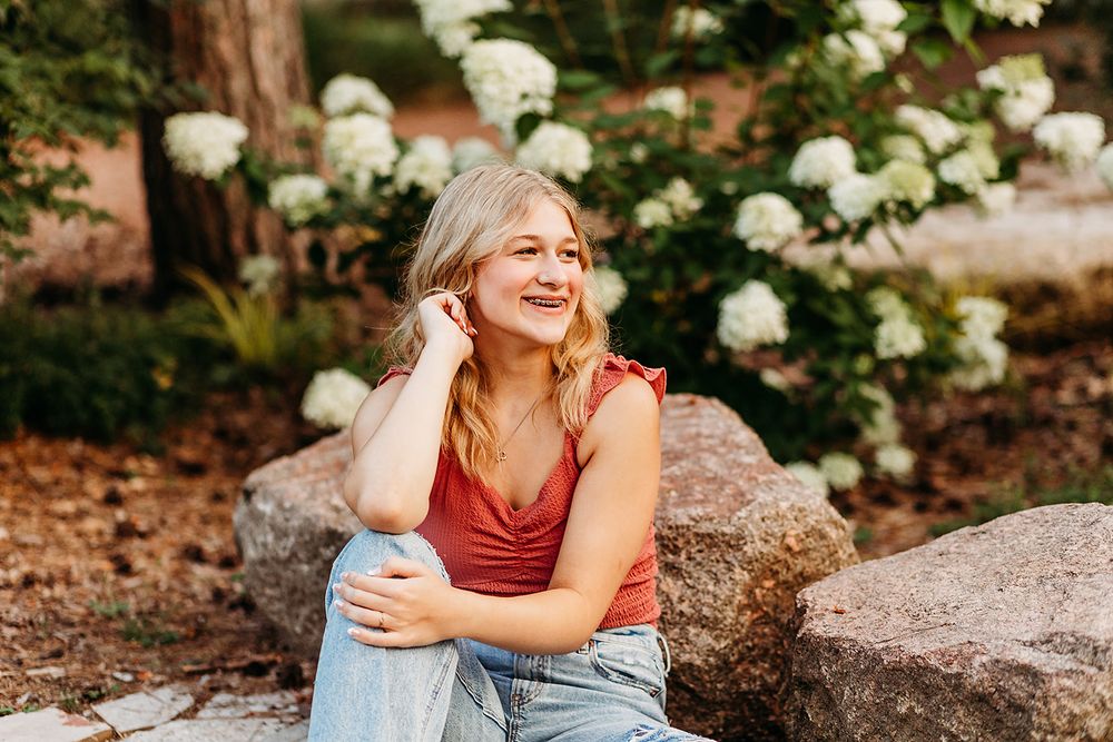 A person in a red tank top sits on rocks in a garden with blooming white hydrangea flowers in the background.