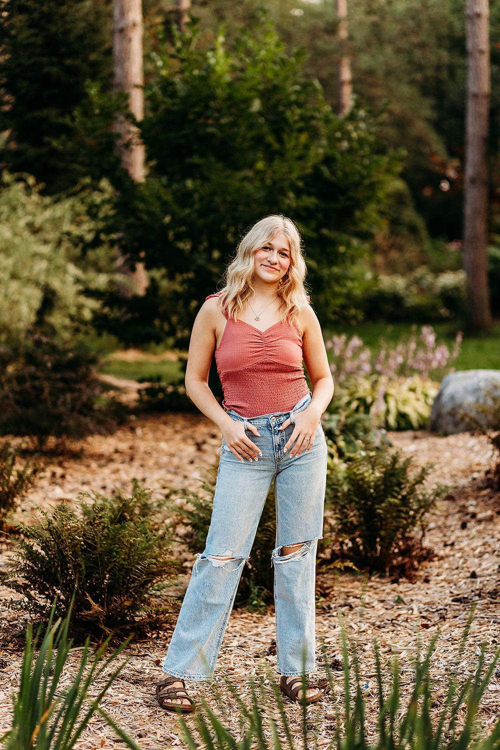 Someone in a coral tank top and distressed jeans poses casually in a garden setting surrounded by evergreen plants.