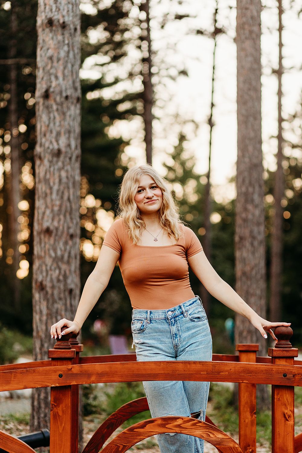 Woman in orange crop top and denim shorts stands smiling on wooden bridge in sunlit forest setting.