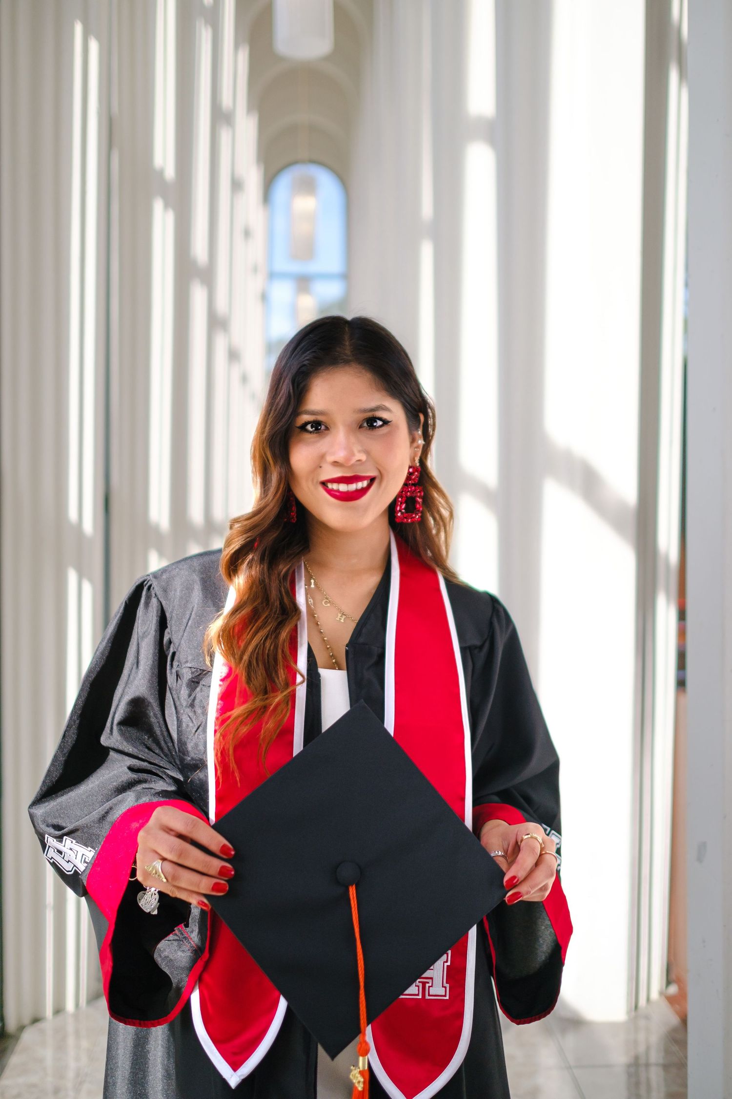 Graduate in cap and gown holding graduation cap while standing in bright hallway with white columns and red stole.
