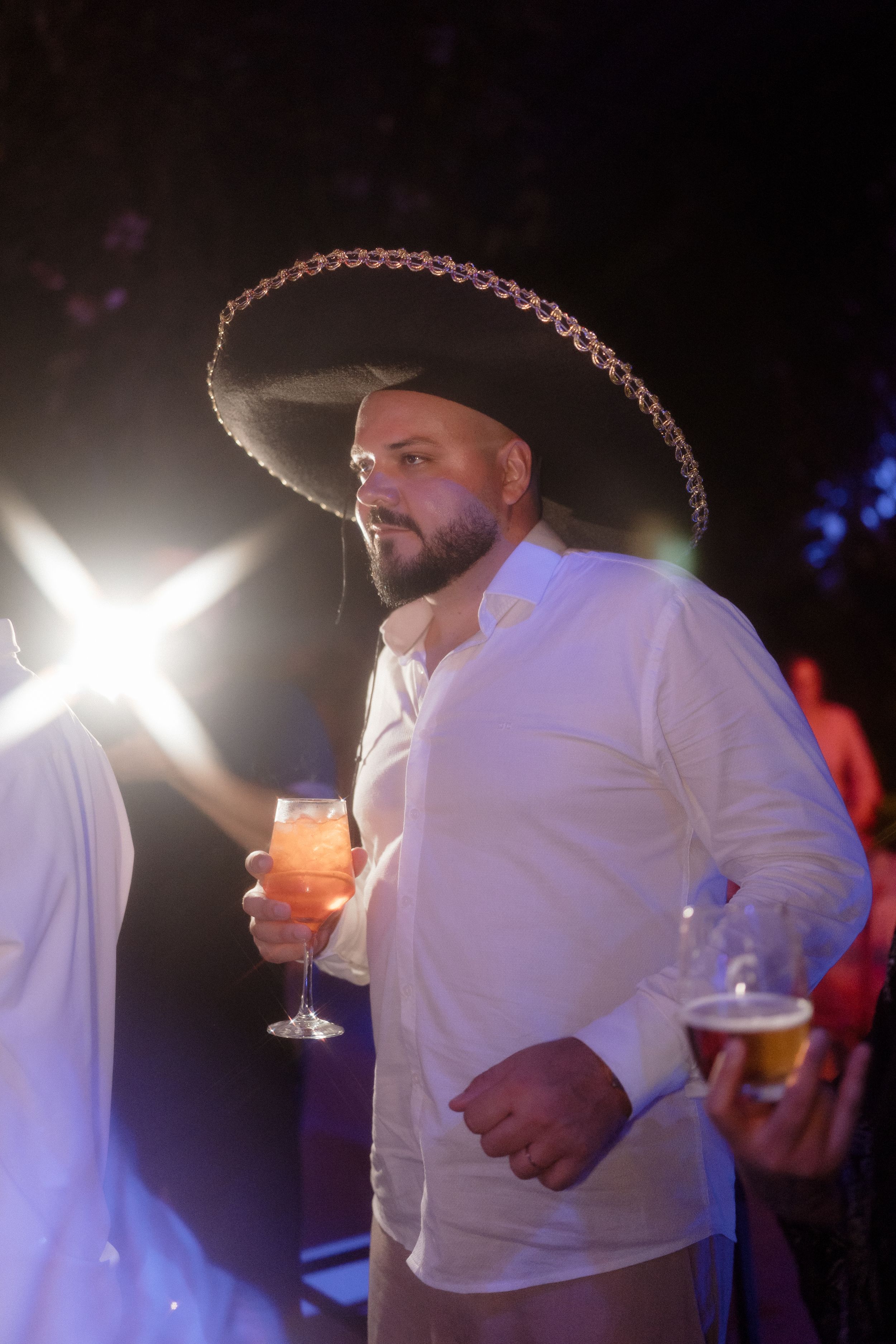 Partygoer in white shirt wearing decorative sombrero while holding cocktail under event lighting.
