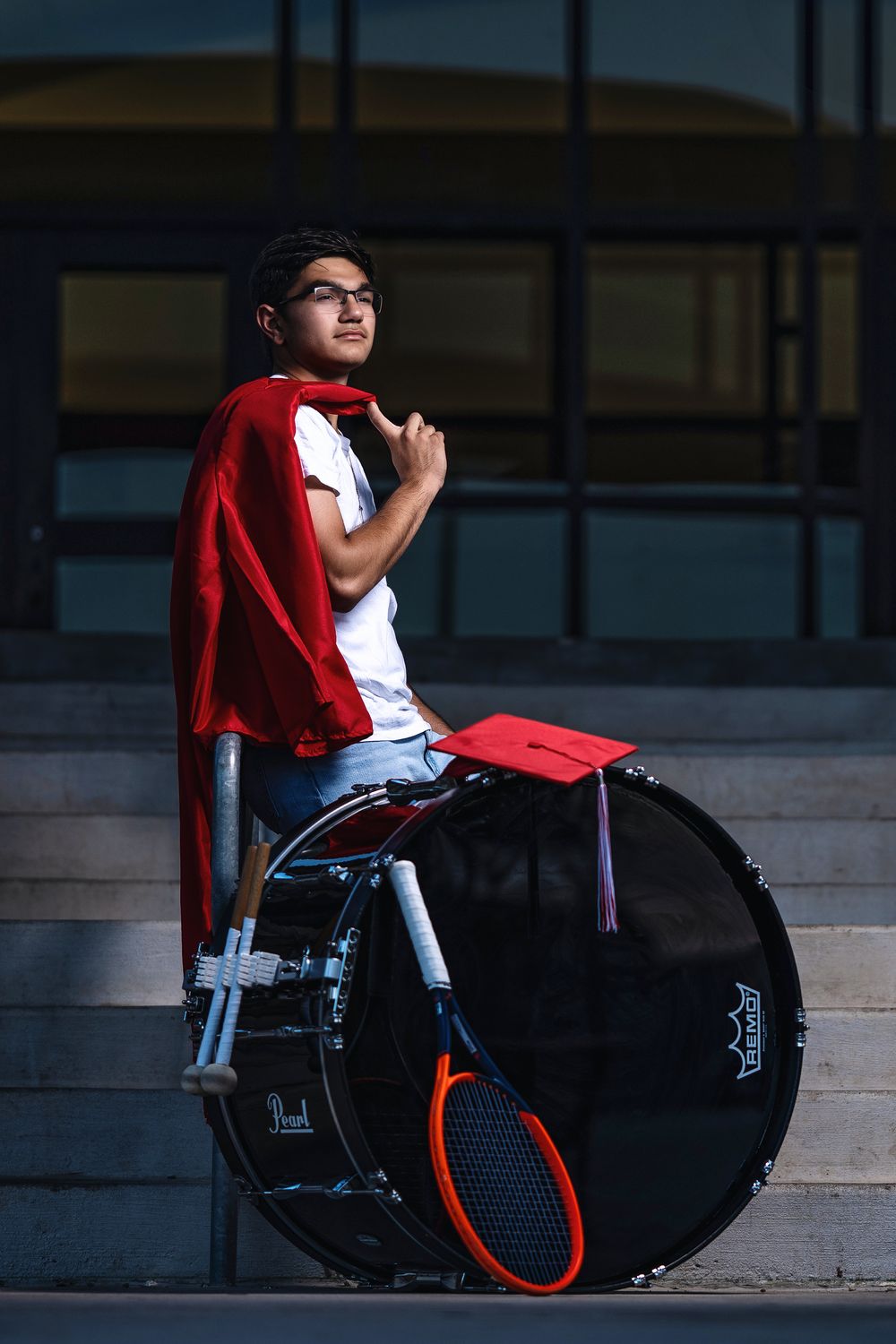 Graduate in wheelchair wearing red cape and graduation cap sits confidently on stairs with large drum in background.
