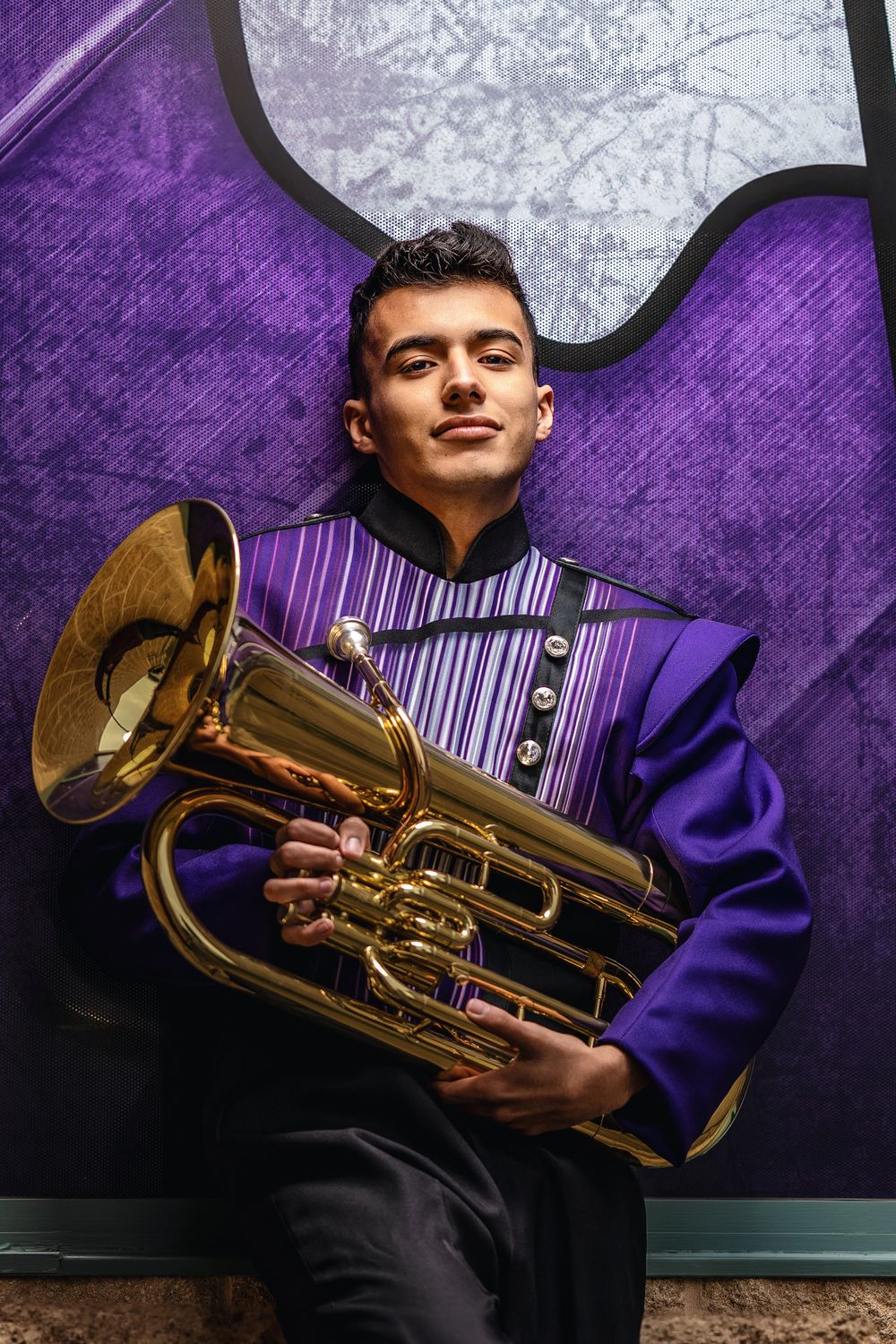 Marching band member in purple uniform holding brass euphonium instrument against purple and gray background.