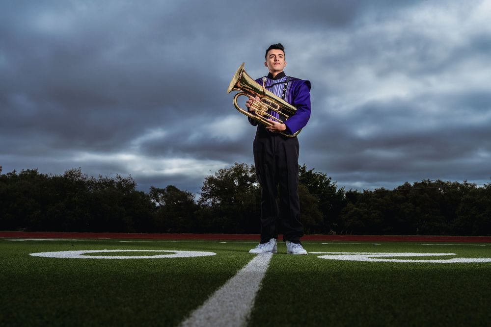 A marching band member stands on an illuminated football field at dusk holding a brass instrument against dramatic clouds.