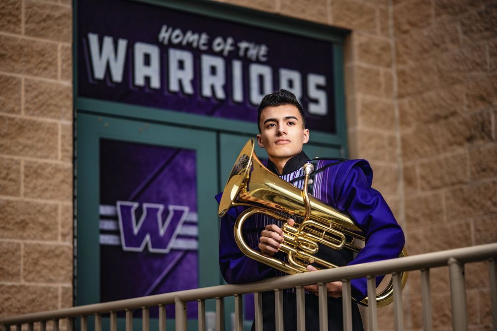 A musician in purple and gold band uniform holds a tuba while standing near a Warriors team logo and brick wall.