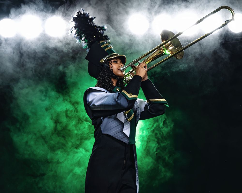 A marching band member plays trombone in uniform against dramatic green smoke and lighting effects.