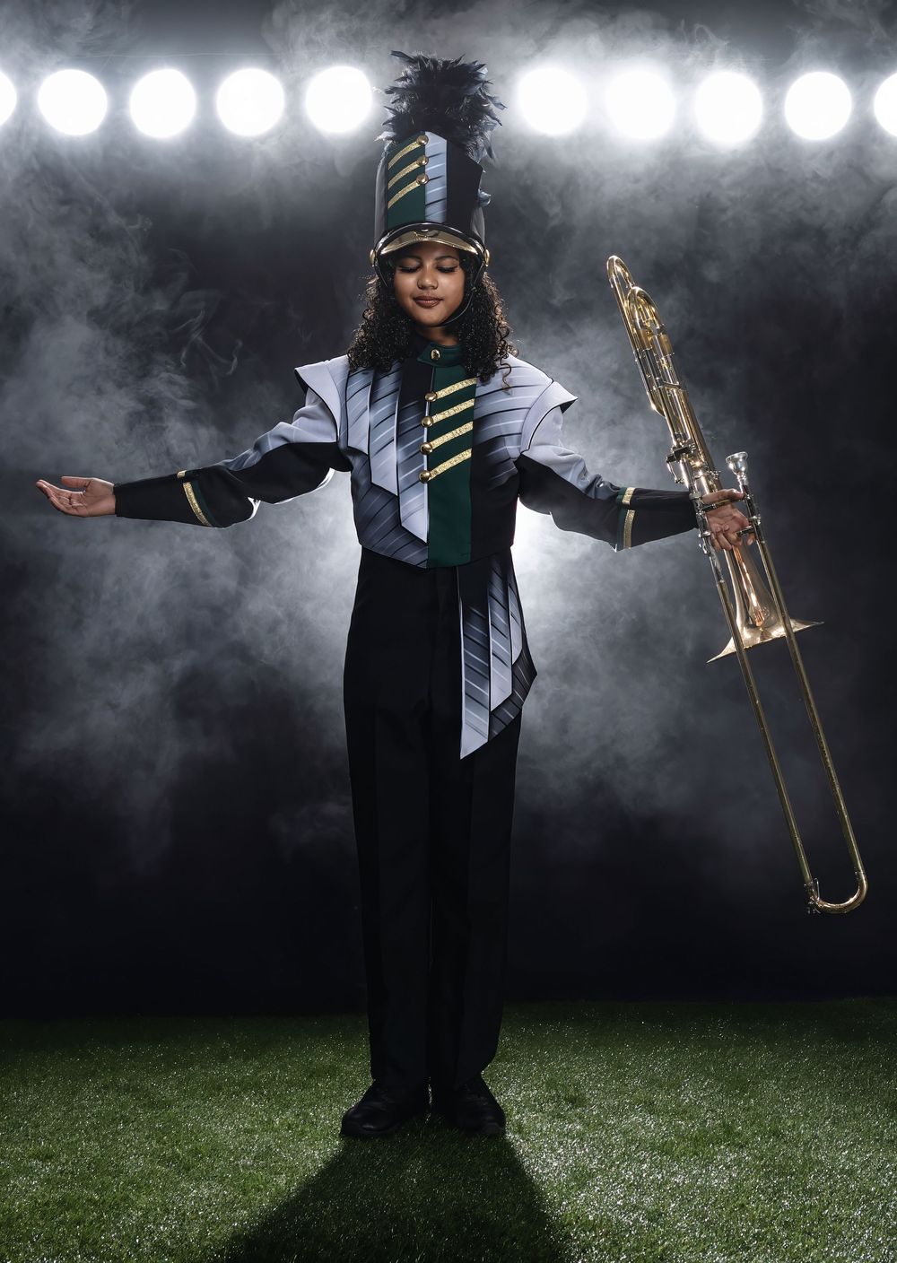 Marching band performer in black and silver uniform with tall plume hat holds a trombone on illuminated field with spotlights.
