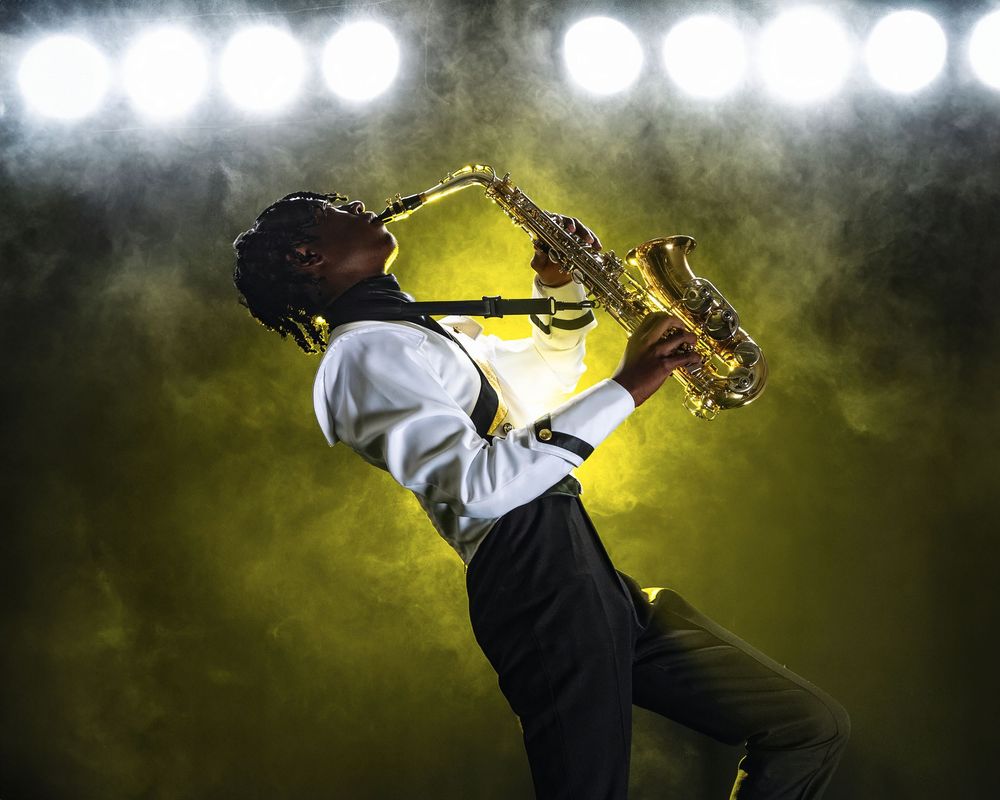 A musician in a white dress shirt plays saxophone under bright stage lights with a dramatic yellow backlight effect.