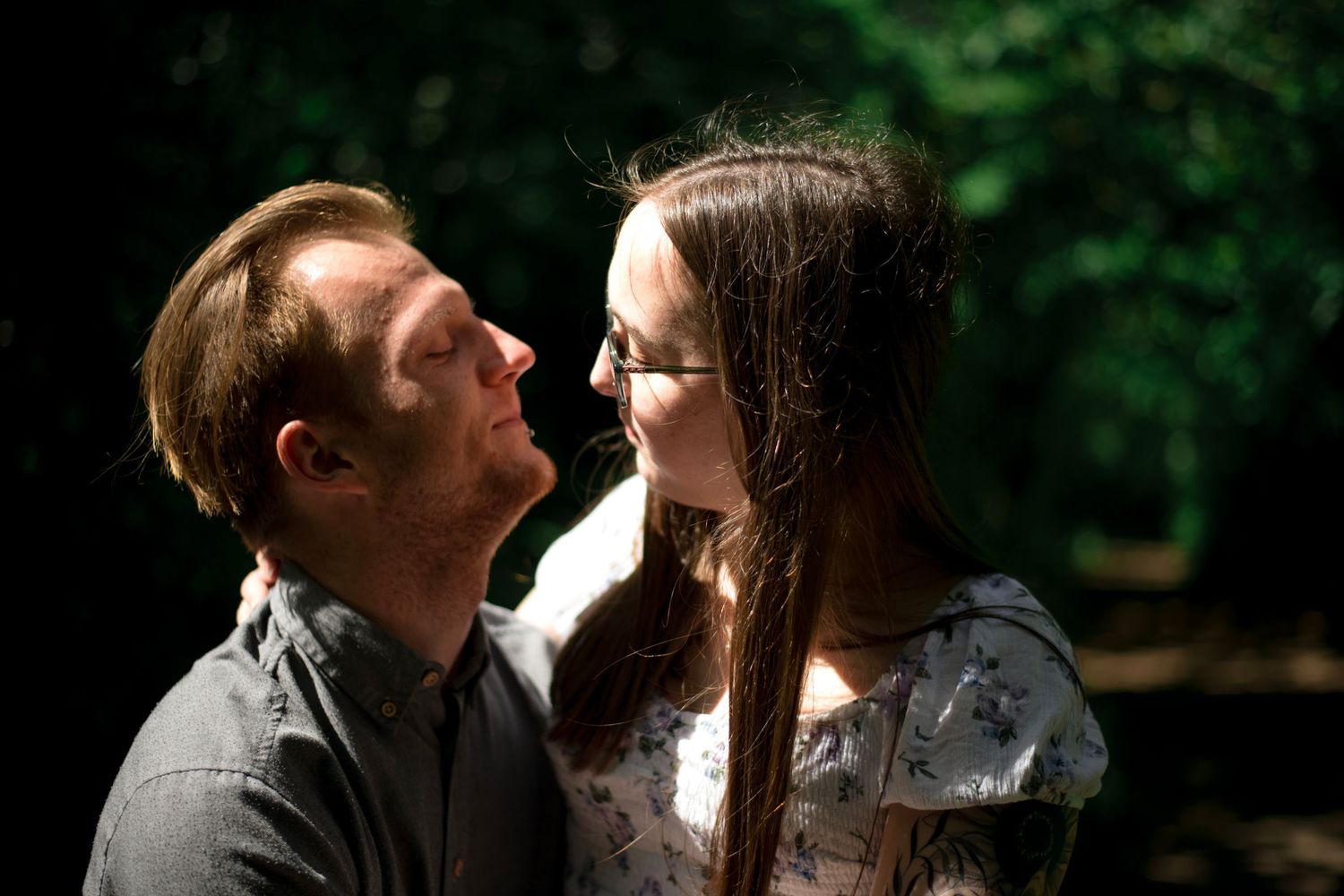 A romantic outdoor couple shares a tender moment while gazing at each other in a sun-dappled forest setting.