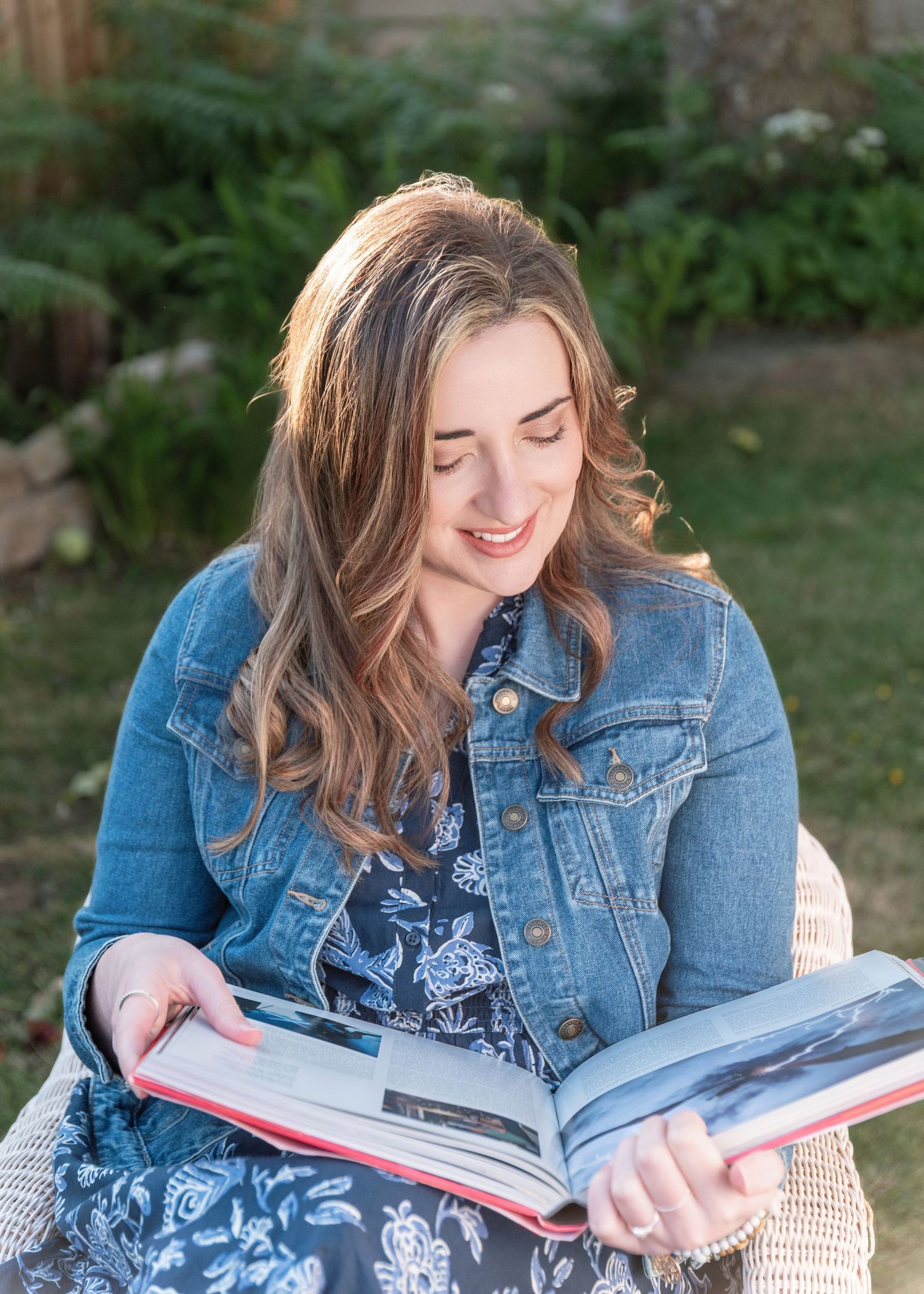 A person in a denim jacket reads a book while sitting outdoors with greenery in the background.