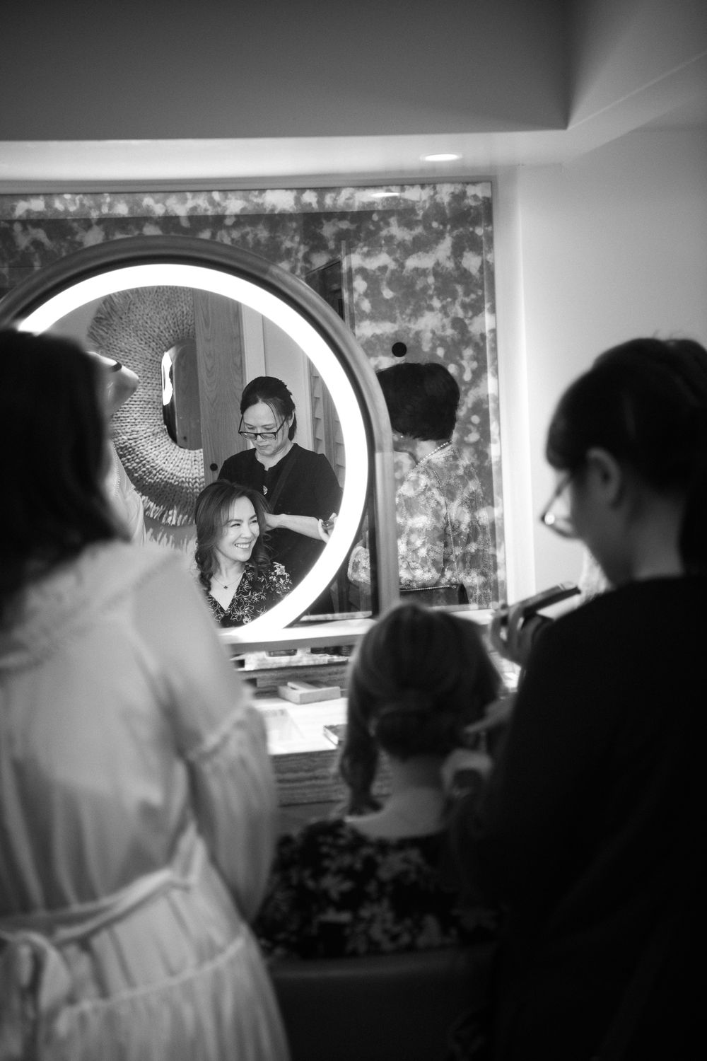 Black and white image showing reflection in round mirror at makeup station while people gather during preparations.