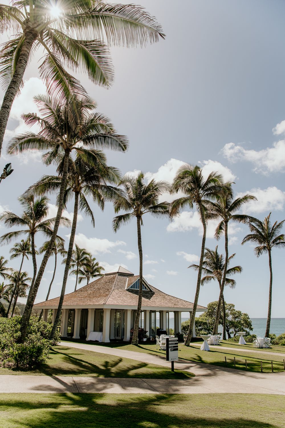 Tropical white pavilion with colonial architecture surrounded by swaying palm trees on a sunny Hawaiian resort oceanfront.