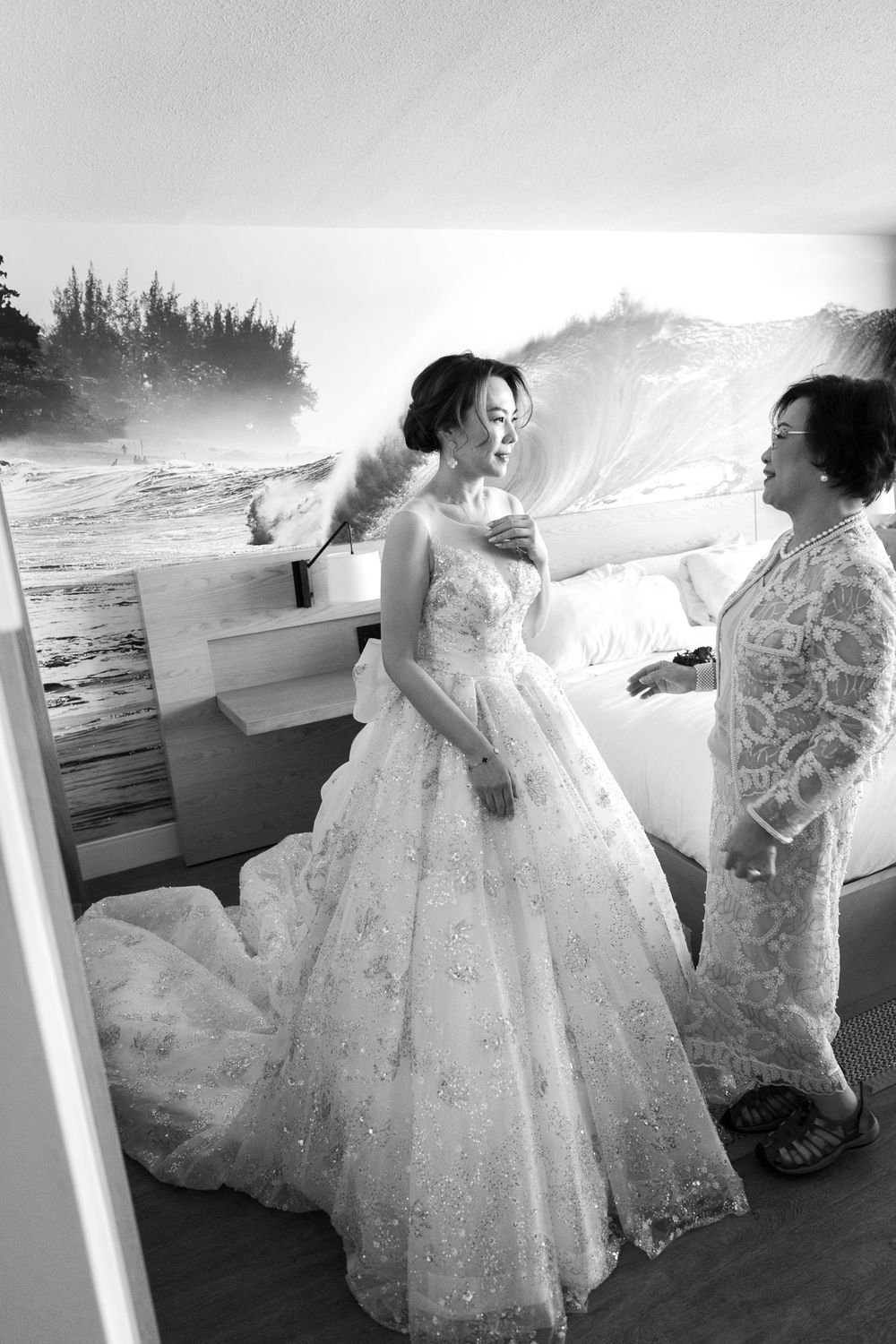 Black and white photo of an elegant bride in a sparkly wedding gown being helped with her dress by the ocean.