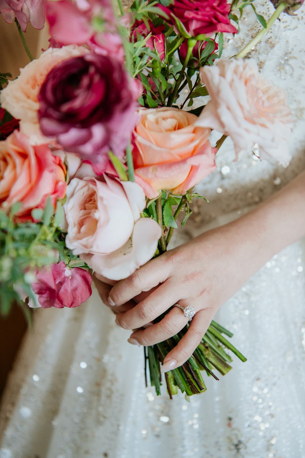 A sparkling wedding gown with delicate pearl details frames a bouquet of pink, peach and burgundy roses tied with ribbon.