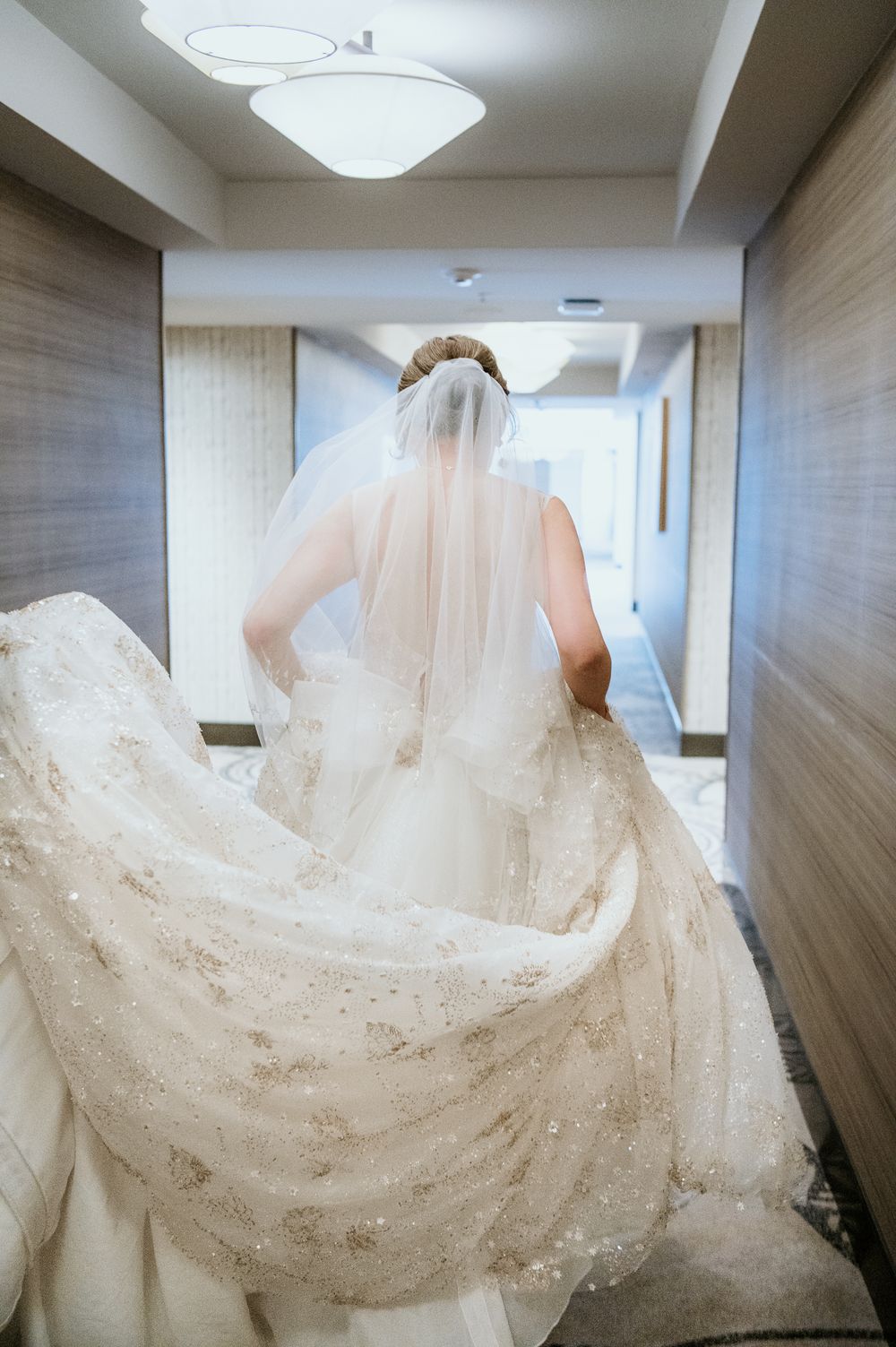 White wedding dress and veil flow elegantly down a modern hallway with wooden walls and overhead lights.