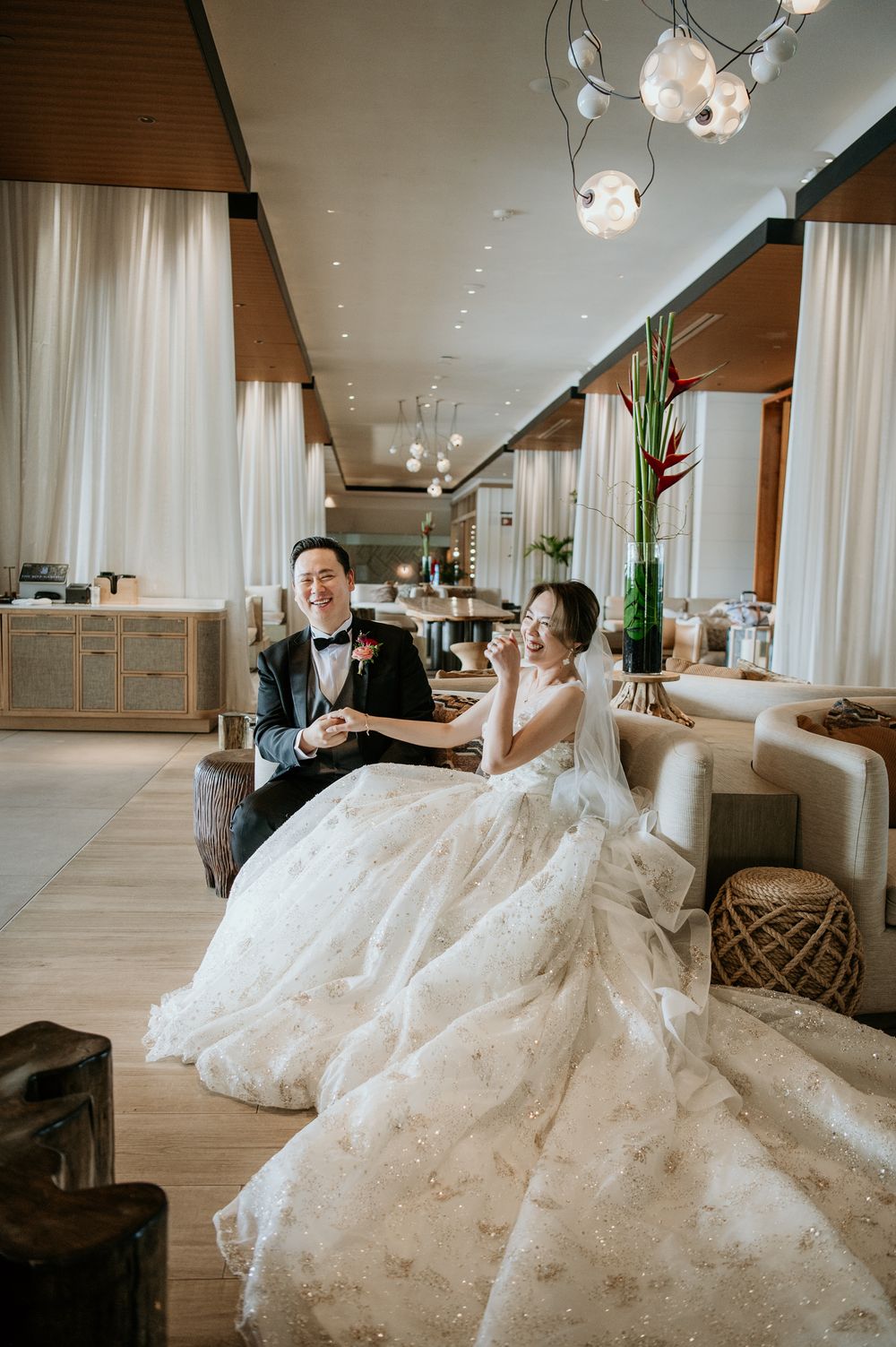 A bride in a flowing white lace gown and groom in tuxedo relax together in an elegant hotel lounge with modern decor.