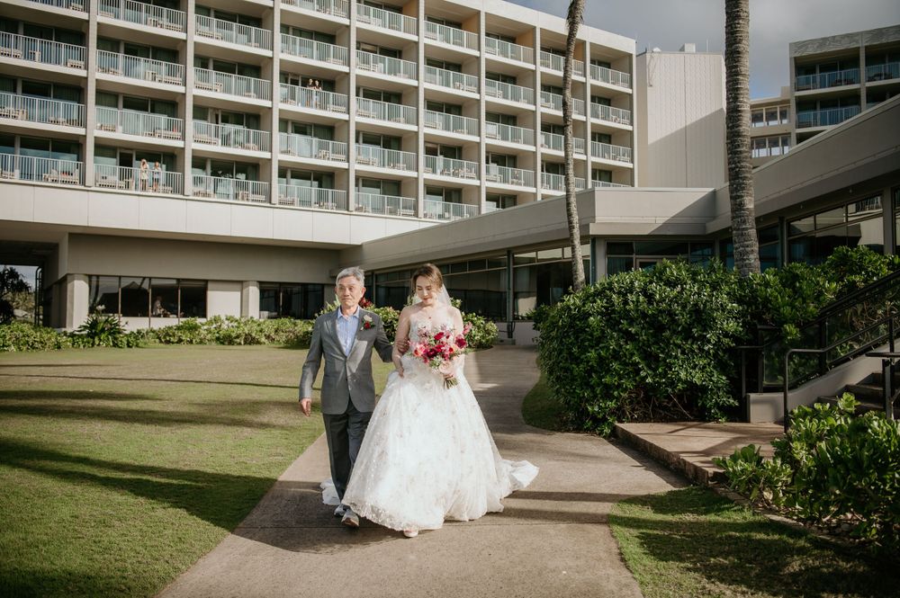 Elegant wedding couple walks together on manicured lawn path outside modern hotel building with balconies.