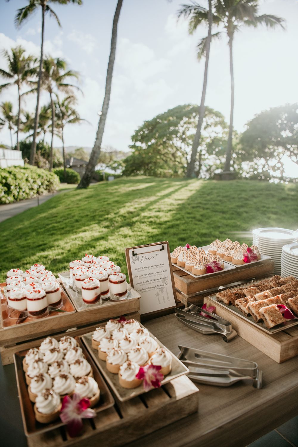 Tropical dessert buffet display with cupcakes and treats on wooden serving trays overlooking palm trees and ocean view.
