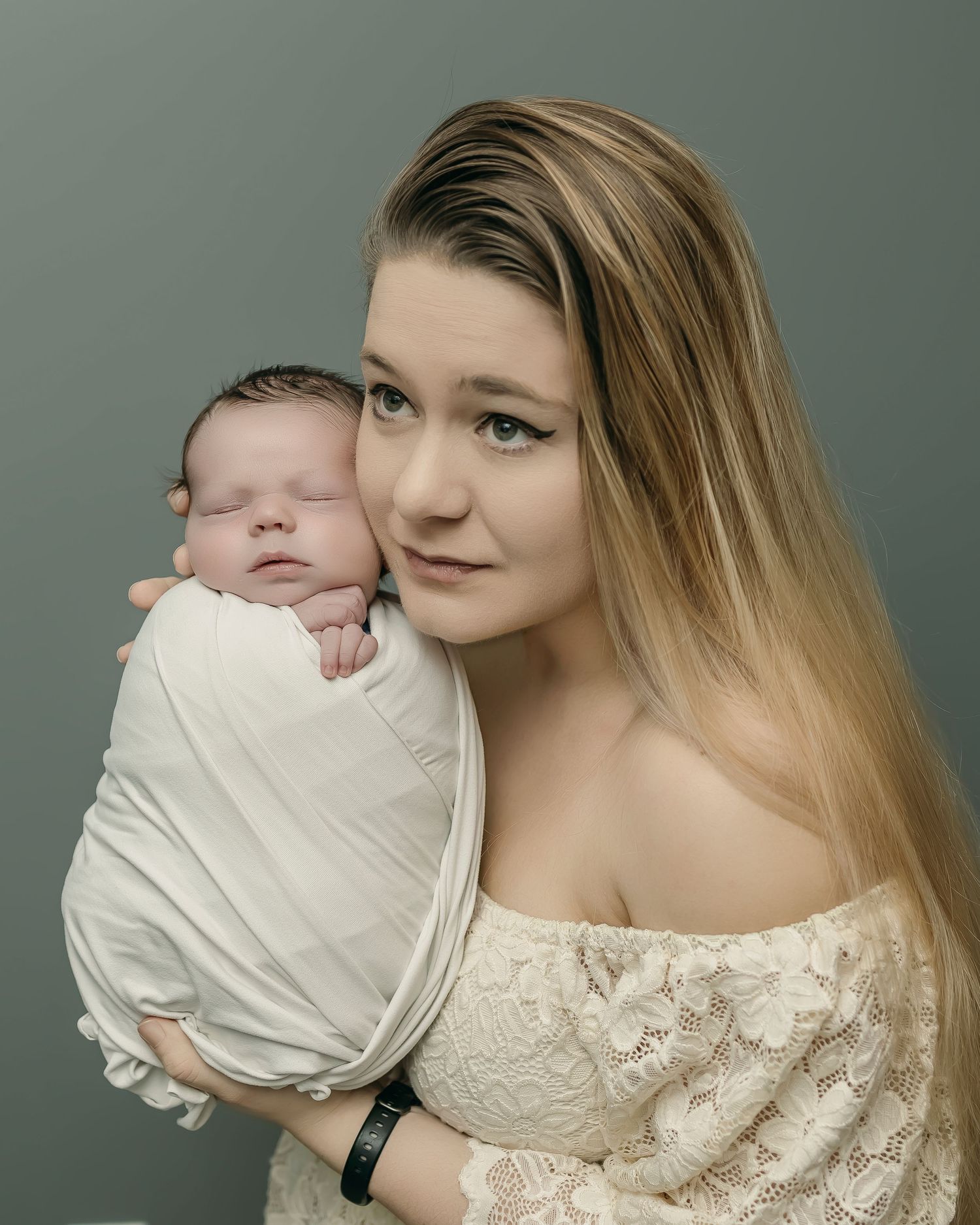 Portrait with newborn baby swaddled in white fabric against grey backdrop, wearing off-shoulder lace dress.