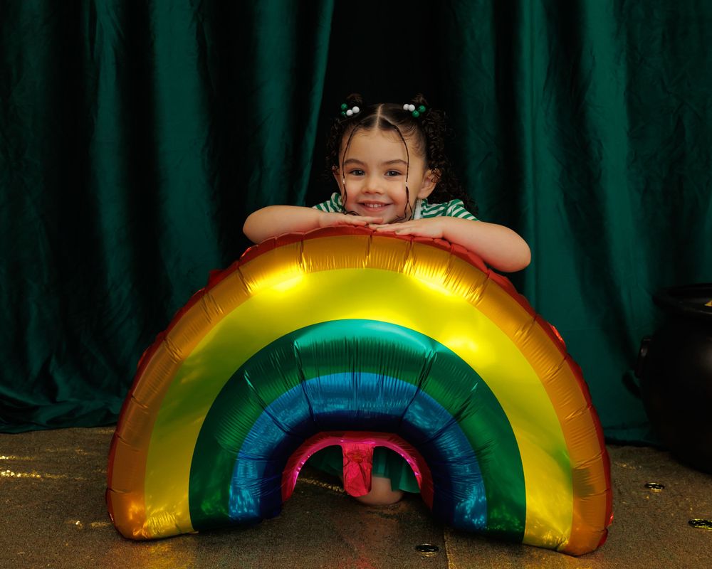 A child poses with a vibrant metallic rainbow balloon prop against a deep green curtain backdrop.