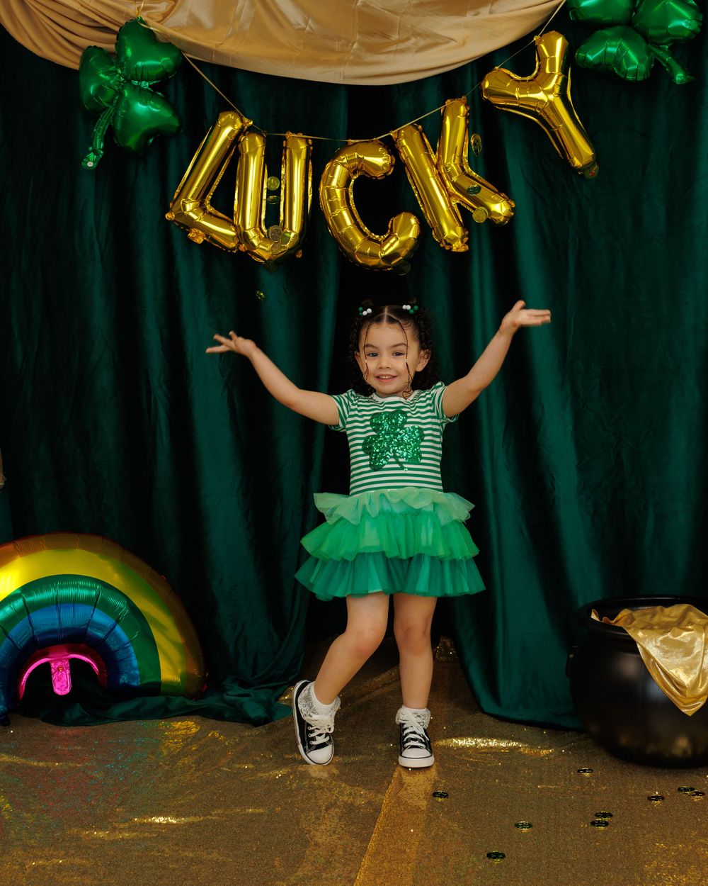 A child in a sparkly green tutu dress poses with arms raised under gold 'LUCKY' balloon letters against a dark green backdrop.