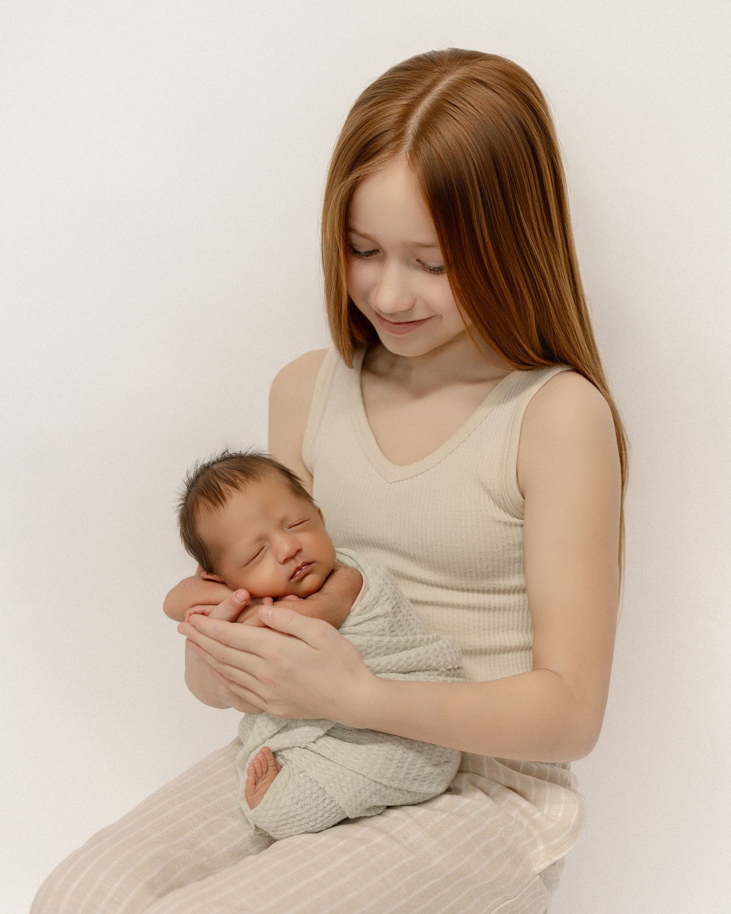 A gentle moment captured as a young woman in a beige tank top tenderly holds and gazes at a sleeping newborn baby.