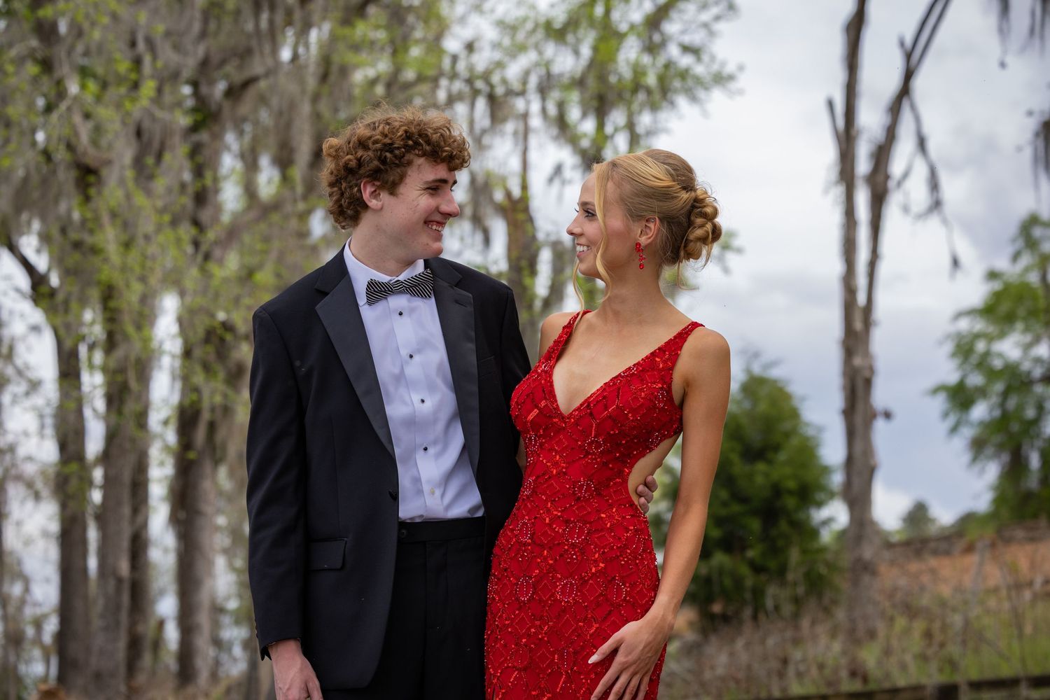 A formal couple poses outdoors in a tuxedo and red gown against trees and Spanish moss.