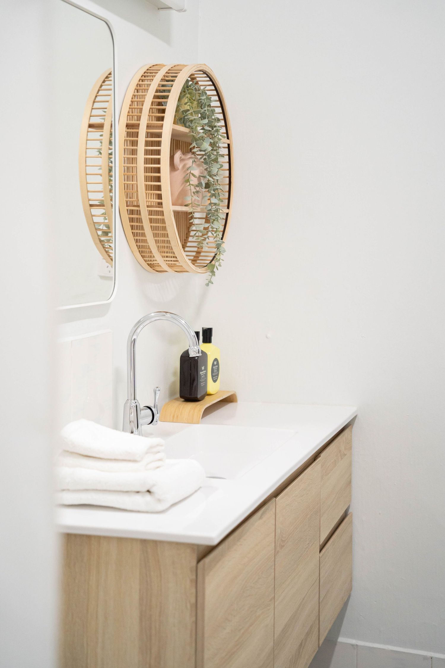 Modern bathroom vanity with wooden cabinets, white countertop, silver faucet, and circular bamboo mirror on white wall.