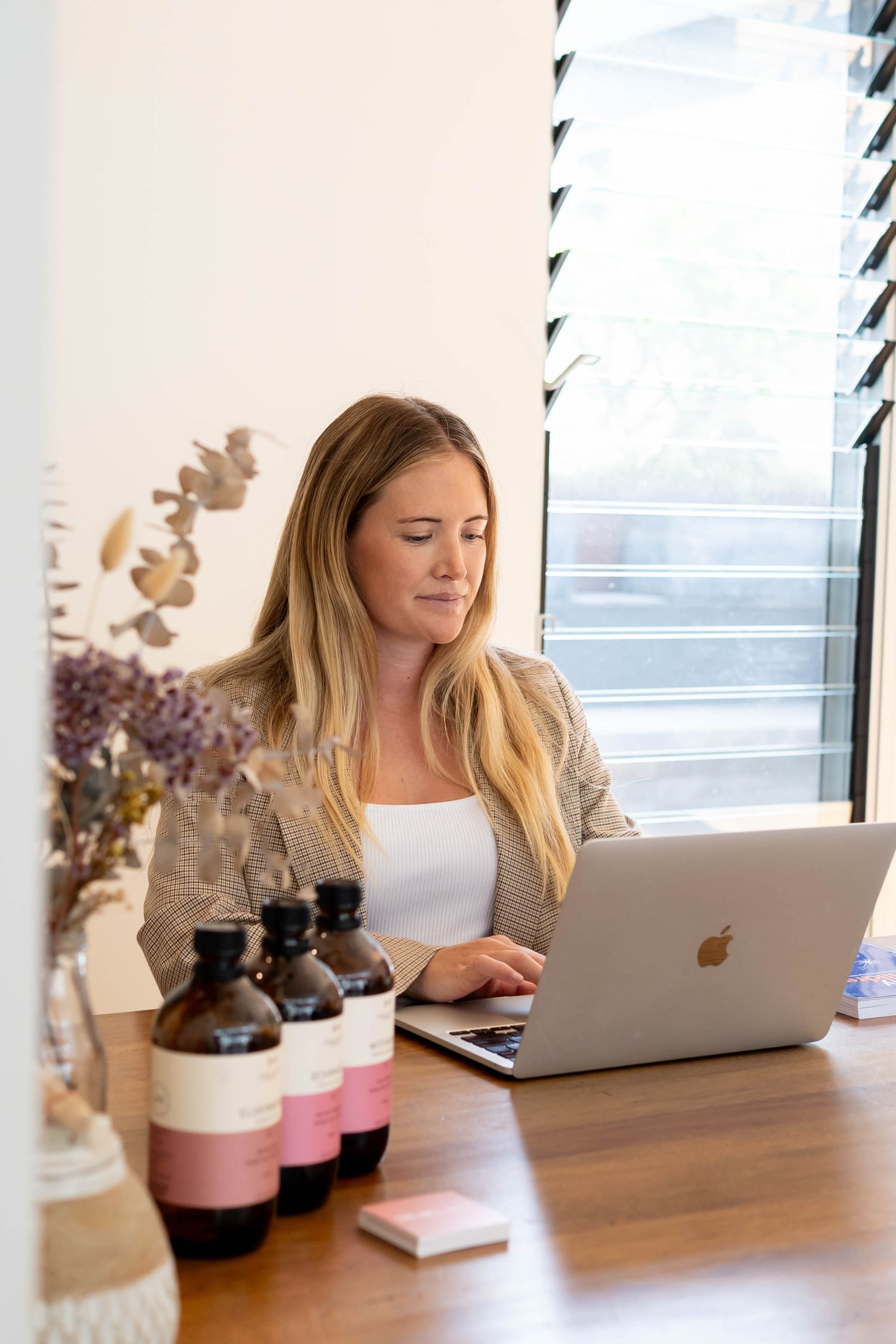 Someone works at a wooden desk with a Macbook laptop while surrounded by pink and brown glass bottles.