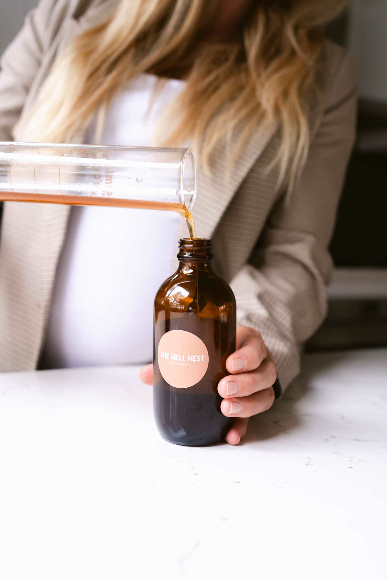 Orange liquid being poured from a glass beaker into an amber apothecary bottle with a round label.