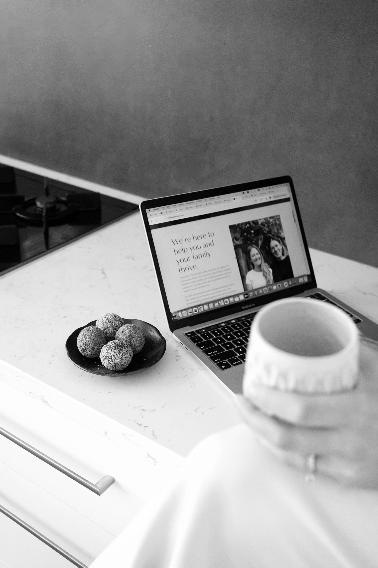 A black and white photo of a laptop, white ceramic cup, and plate of cookies on a white desk surface.