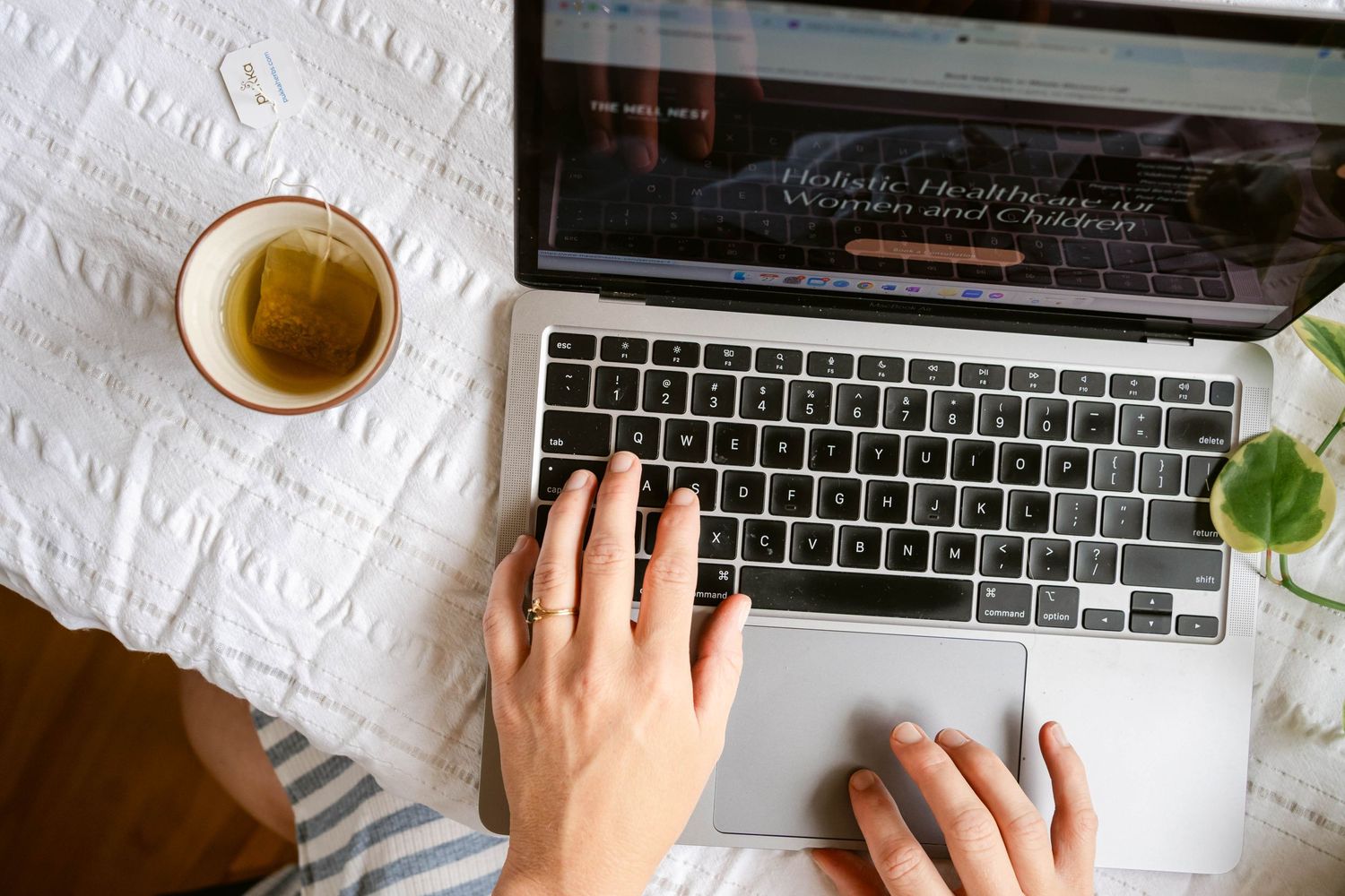 A cup of tea sits beside hands typing on a MacBook laptop keyboard with a small plant leaf in the corner.