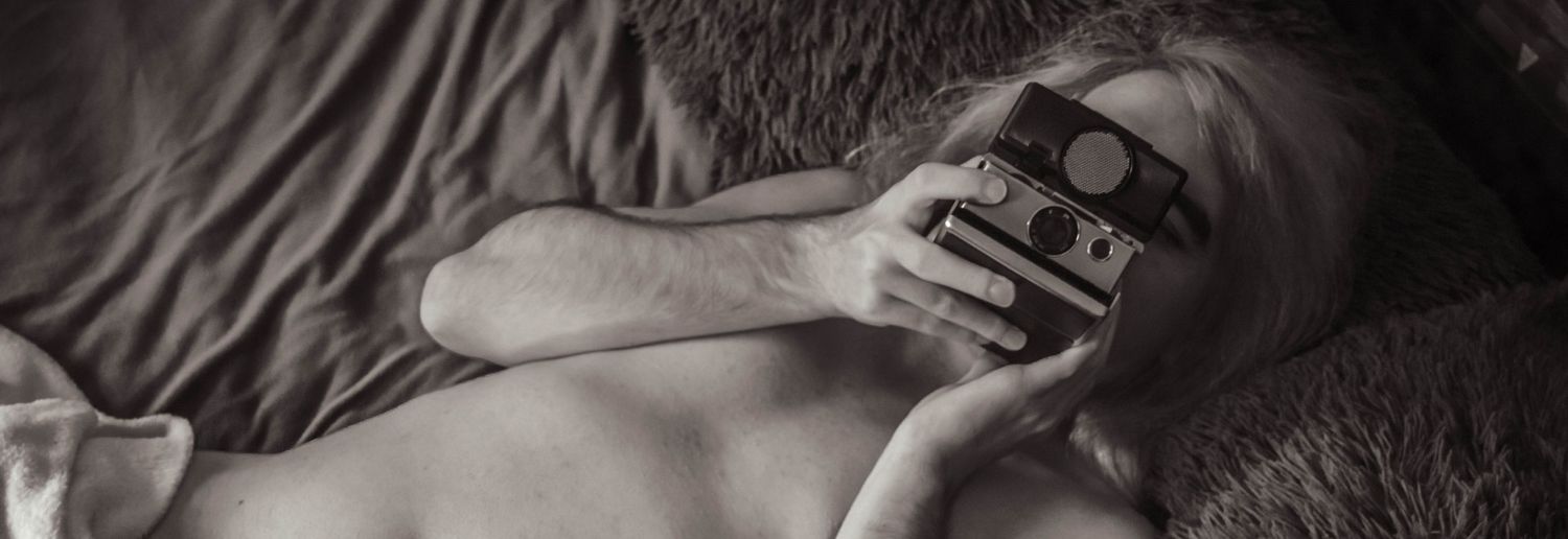A black and white photo of a person lying on a bed mostly nude with an old school polaroid camera