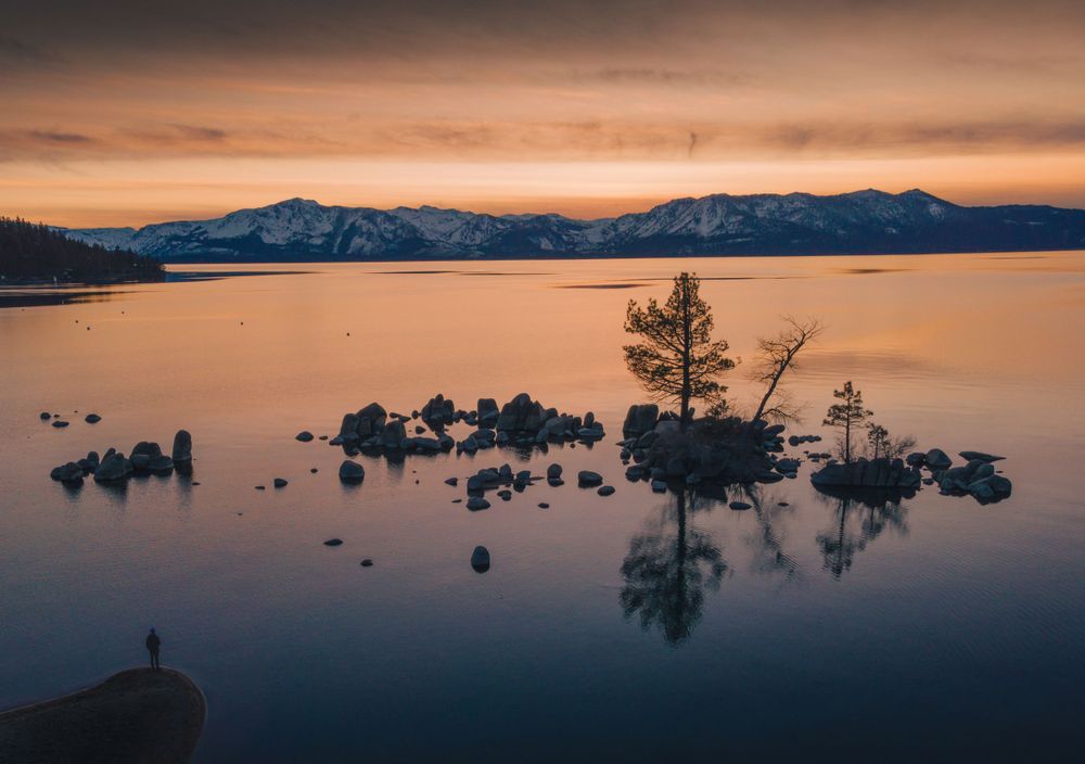 A lone tree stands silhouetted on a rocky outcrop during sunset at Lake Tahoe with snow-capped mountains in the distance.