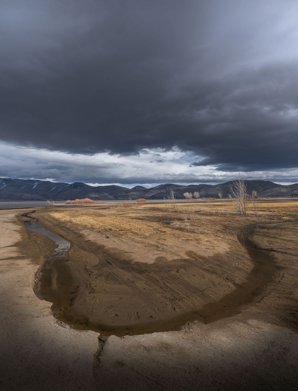 Dramatic storm clouds loom over a winding dirt path through arid terrain with mountains in the distance under moody skies.