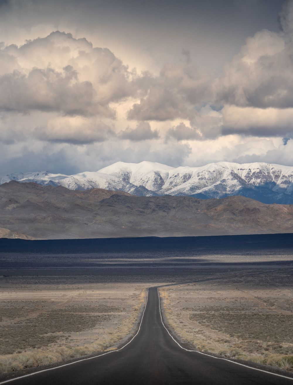 A dramatic mountain road stretches toward snow-capped peaks under stormy clouds in the Nevada desert landscape.