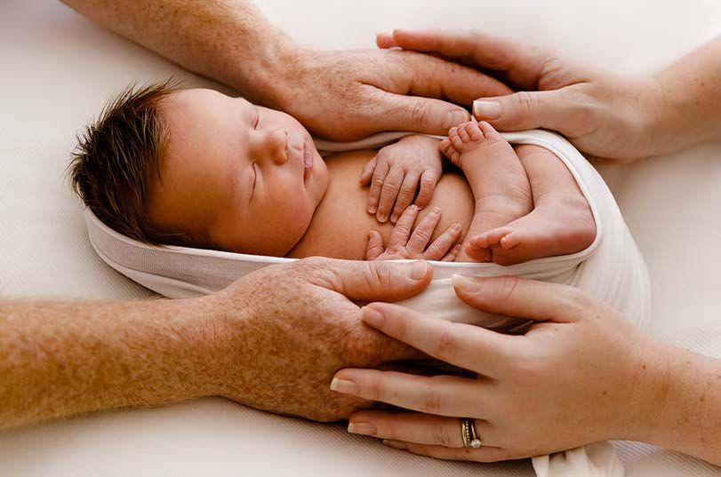 Adult hands gently cradle a sleeping newborn baby wrapped in soft white cloth against a light background.