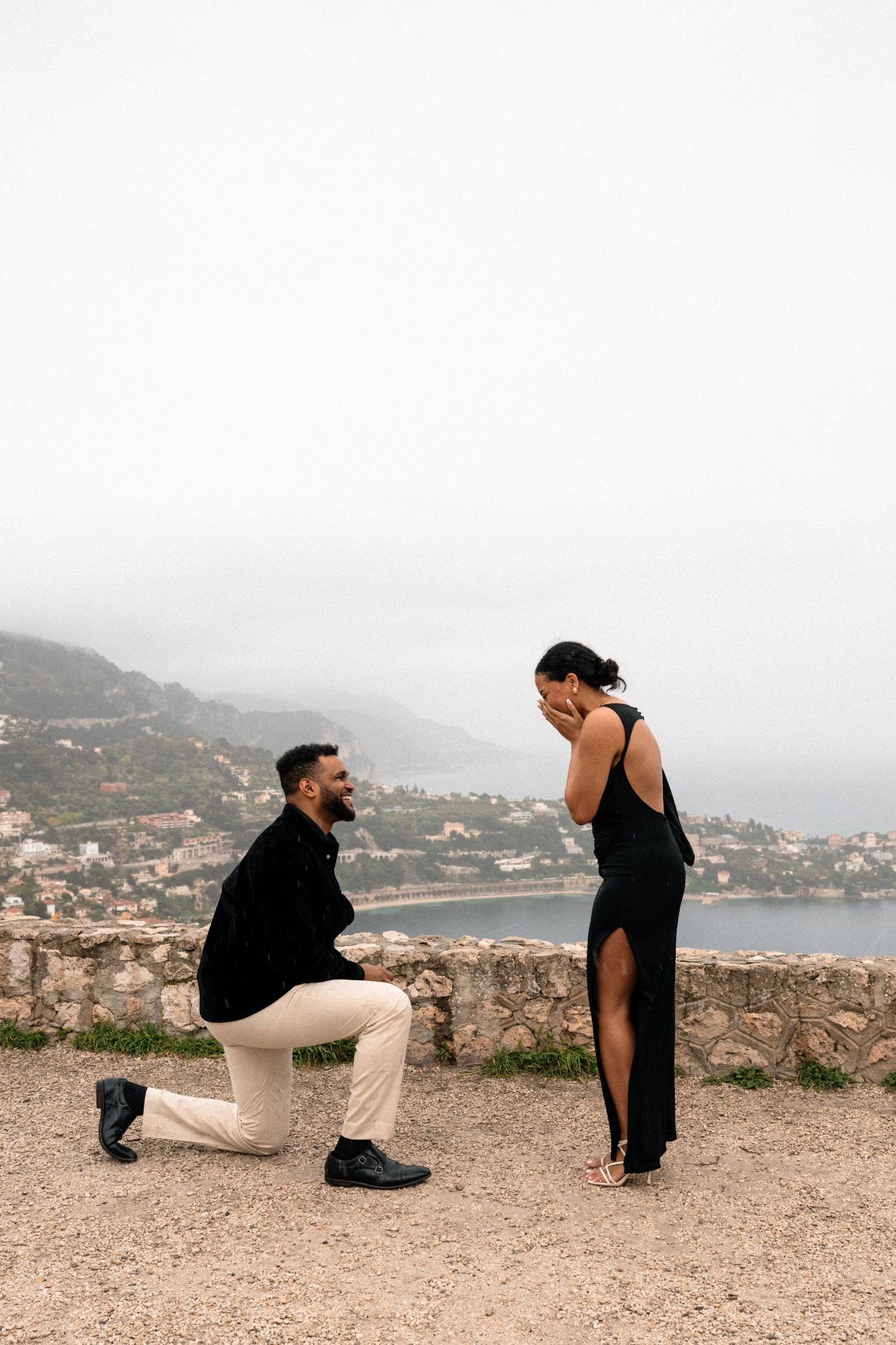 A romantic proposal scene on a scenic overlook with coastal mountains and water in the background.