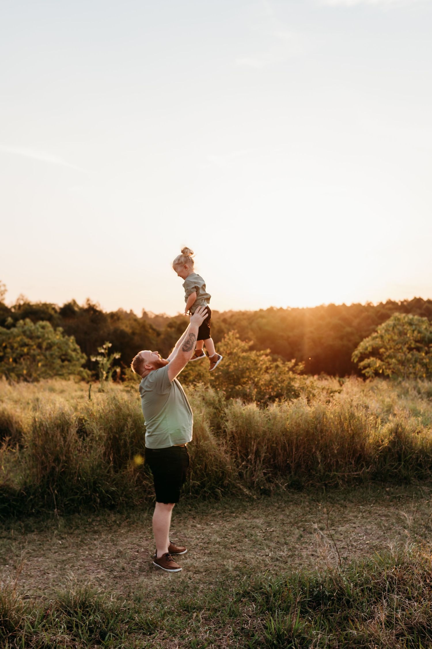 Parent lifting child playfully in air against sunset sky in natural field setting.