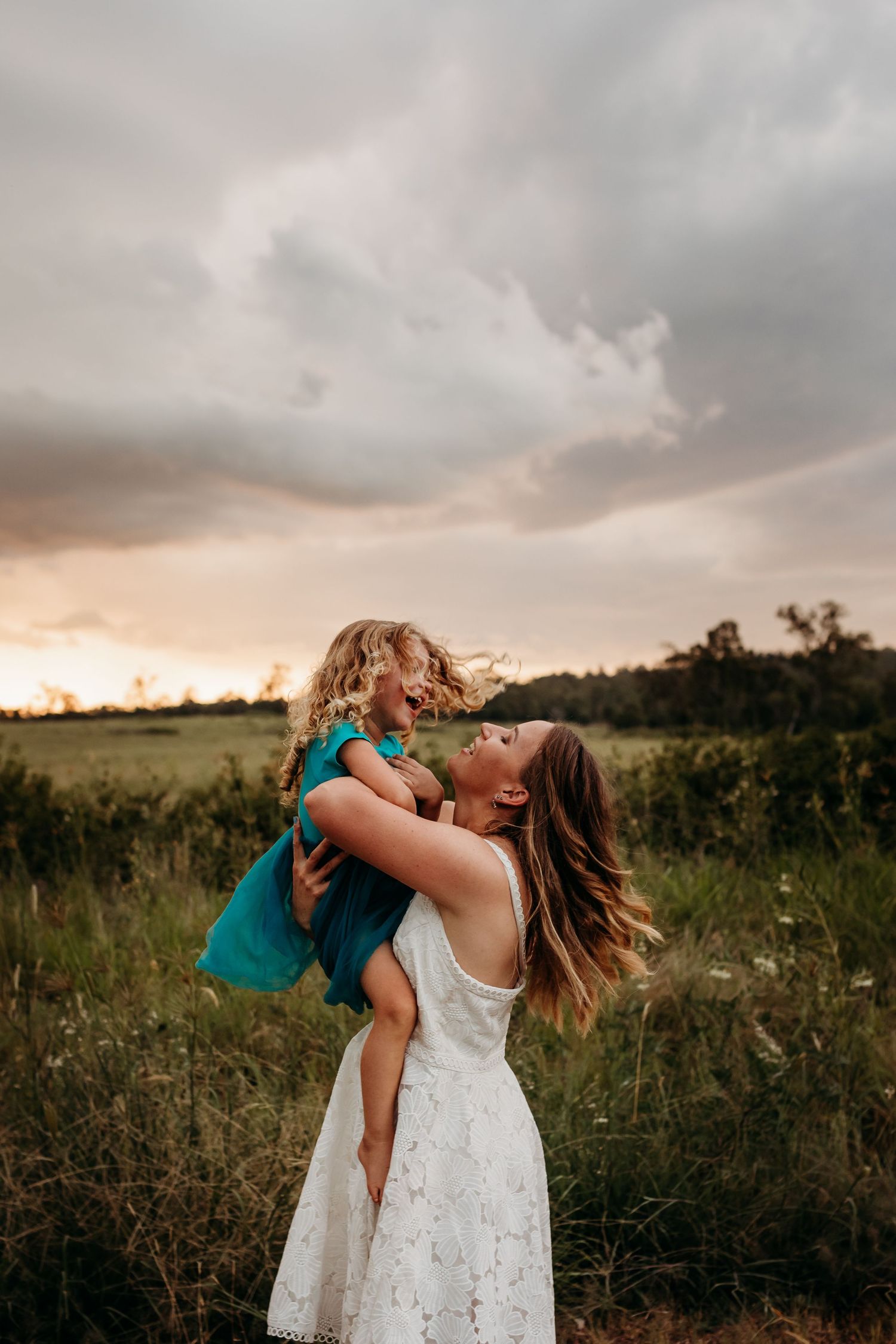 Mother and child playing in field during golden hour with dramatic cloudy sky.