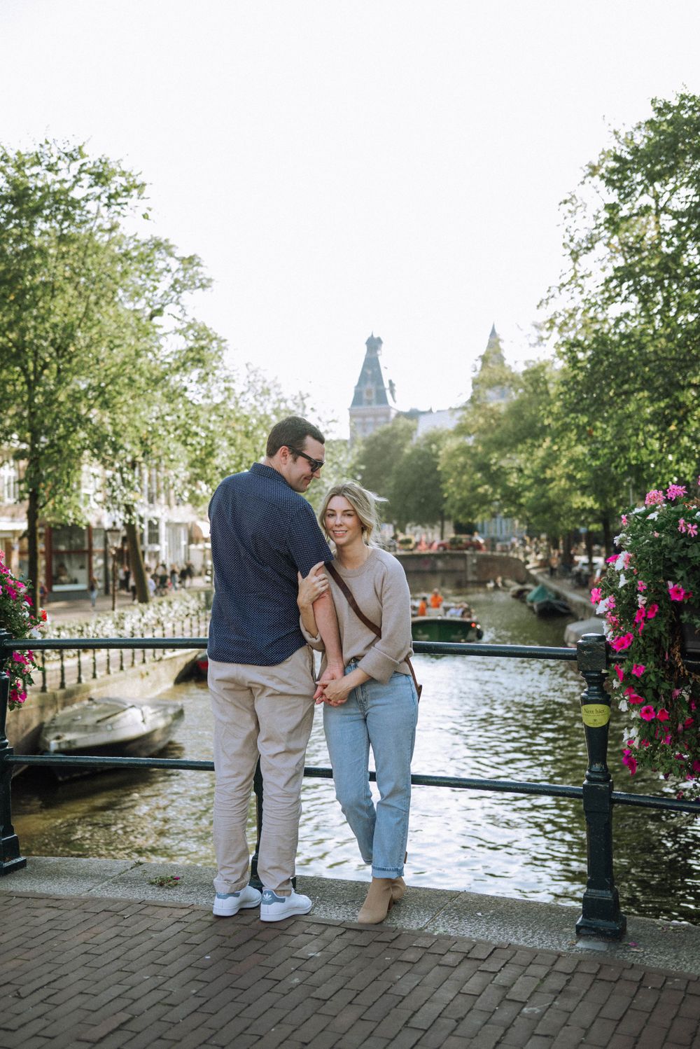 A couple stands on a cobblestone path along a canal with flowers and boats in Amsterdam.